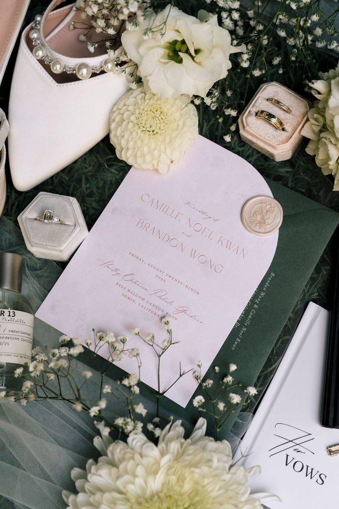 Flat lay of invitation, vow book, bridal shoes and white dahlias on green