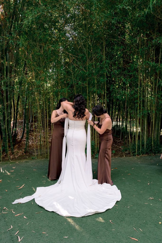 Bridesmaids fastening the bride's gown in the bamboo garden at Hartley Botanica