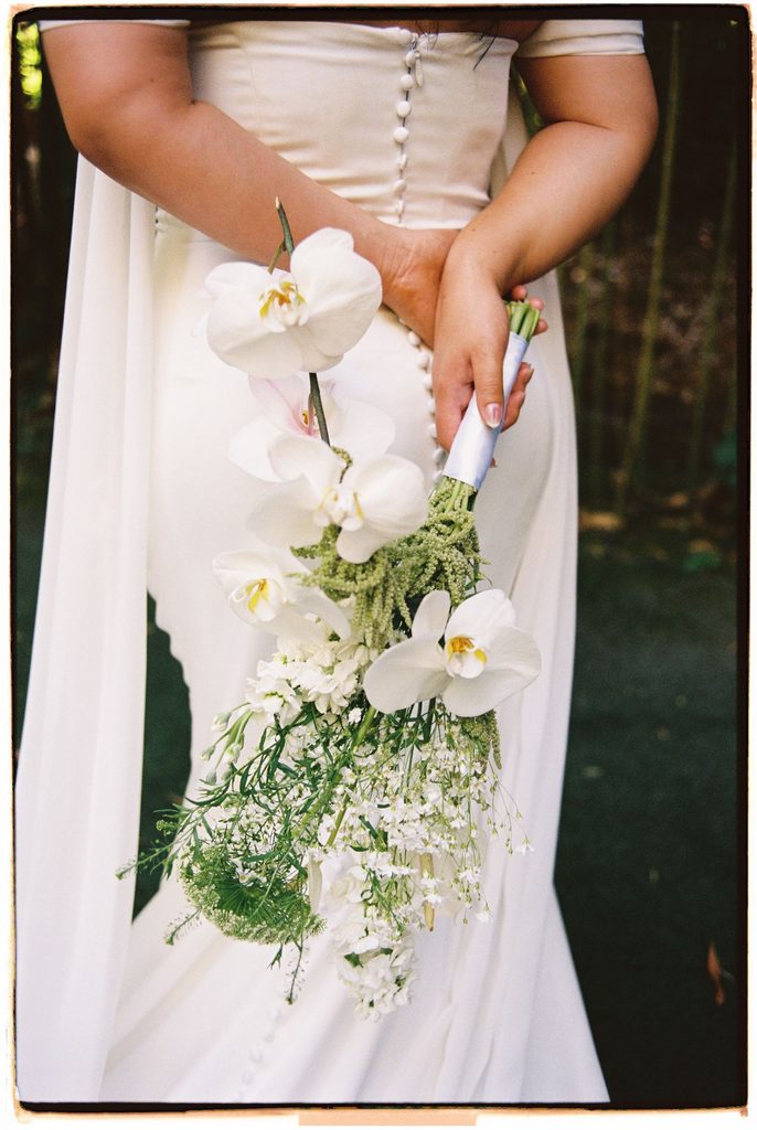 Bride's hands holding a white orchid bouquet behind her back, close up