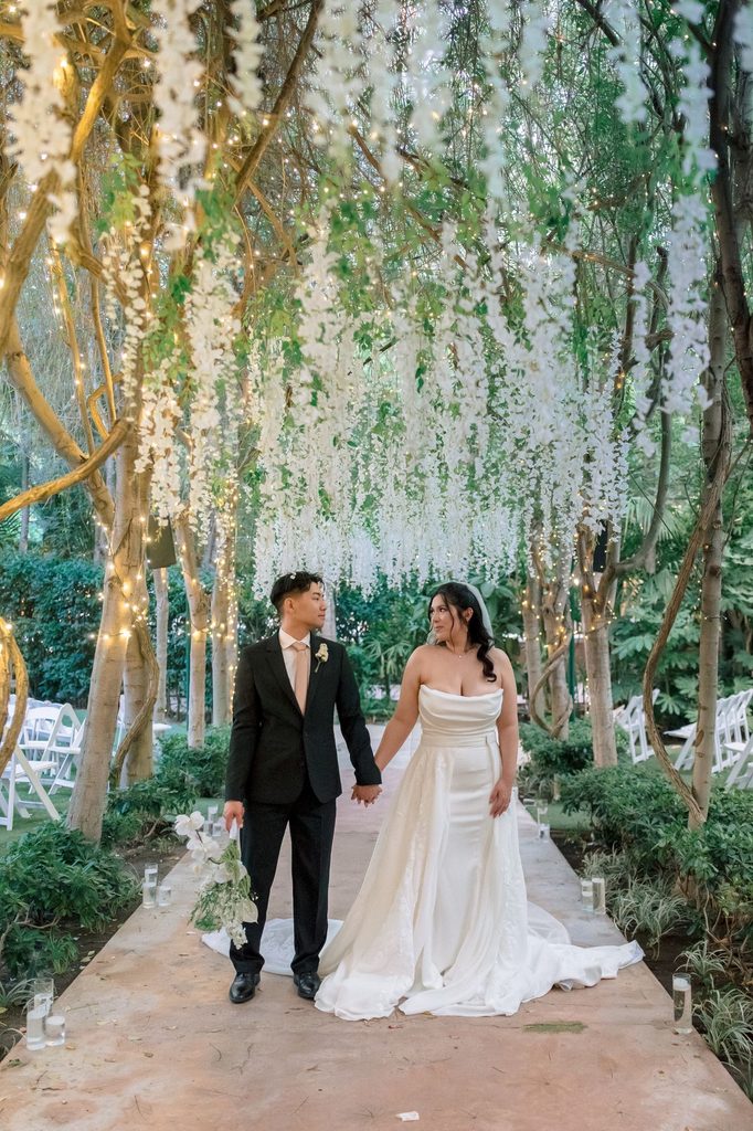 Bride and groom walking hand in hand beneath the hanging blooms